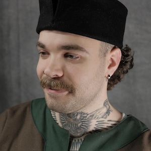 Op de afbeelding: Een zwarte wollen hoed met een V-vormig detail aan de voorkant. De hoed wordt gedragen door een persoon met een snor en een tatoeage op zijn nek. De persoon draagt een bruin en groen shirt. De afbeelding bevat het "FREYHAND" logo.