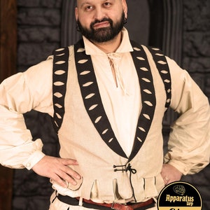 Pode incluir: Um colete de linho bege com guarnições e recortes pretos. O colete tem um fecho de gravata e é usado sobre uma camisa branca. A pessoa que usa o colete tem barba e está em pé em frente a uma parede de pedra.