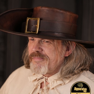 May include: A man wearing a brown leather hat with a gold buckle and a white shirt with a tied collar. The hat has a wide brim and a tall crown. The man has a long beard and mustache. The shirt has a ruffled collar. The man is looking to the side. The image has a text overlay that says "Apparatus Larp Shop".
