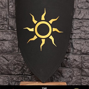 Op de afbeelding: Een zwart schild met een gouden zonontwerp. Het schild is gemaakt van schuim en is een replica van het schild dat door de Witcher Geralt van Rivia wordt gebruikt in de videogame The Witcher 3: Wild Hunt. Het schild wordt getoond op een houten standaard met de tekst "The Witcher" en het CD Projekt Red-logo.