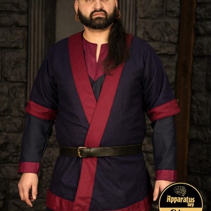 Peut inclure: Une tunique bleu foncé avec une bordure bordeaux et une ceinture en cuir noir. La tunique a un col en V et des manches longues. Le modèle porte une ceinture en cuir noir avec une boucle en laiton.