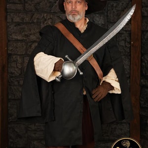 May include: A person wearing a black coat and brown pants holds a silver sword with a round pommel. The person is wearing a brown leather belt and a brown hat with a buckle. The background is a stone wall.