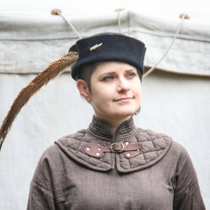 Può includere: Una donna indossa una giacca trapuntata marrone con un cinturino e una fibbia in pelle. Indossa un cappello nero con una piuma attaccata. Il cappello ha uno spillo dorato sul davanti.