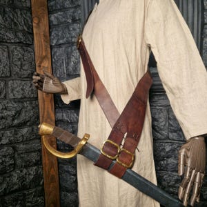 May include: A brown leather sword scabbard with two brass buckles. The scabbard is worn diagonally across the body and secured with a strap.