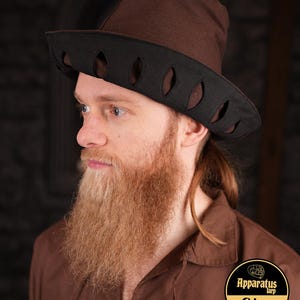 May include: A brown felt hat with a black brim and cut-out design. The hat has a tall crown and a wide brim. The hat is worn by a person with a long beard.