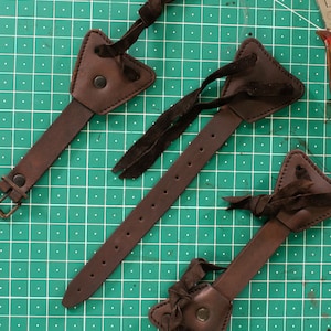 Może przedstawiać: Trzy brązowe skórzane paski z trójkątnymi skórzanymi elementami na końcach. Paski mają dziurki wybite wzdłuż długości i są wiązane brązowymi skórzanymi sznurkami.