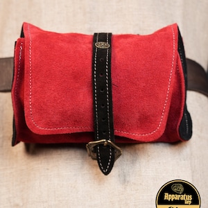 May include: A red suede pouch with a black leather strap and a brass buckle. The pouch has white stitching around the edges.