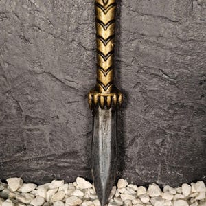 May include: A decorative dagger with a gold-colored handle and a dark blade. The handle features a star-shaped design at the top and a chevron pattern. The dagger is set against a textured dark gray background.