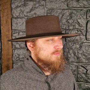 May include: A brown hat with a flat top and wide brim. The hat is made of a felt-like material and has a dark brown band around the base of the crown. The hat is worn by a person.