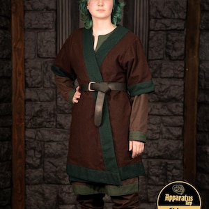 Puede incluir: Una túnica marrón con ribetes verdes y un cinturón negro. La túnica tiene cuello en V y mangas largas. La persona que lleva la túnica tiene el pelo verde y está de pie frente a una pared de piedra.