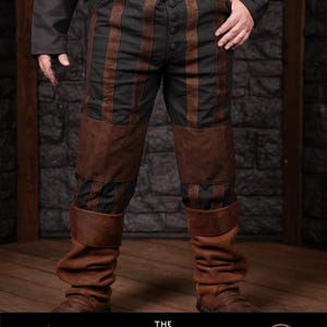 May include: Black and brown striped trousers with leather accents. The trousers have a high waist and are cinched with a brown belt. The trousers are worn by a person standing on a wooden floor.