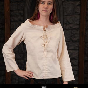 Op de afbeelding: Een wit, lang mouwig shirt met een veterdecolleté. Het shirt heeft een eenvoudig ontwerp en is gemaakt van een lichtgewicht stof. Het shirt is gelabeld met "The Witcher" en het CD Projekt Red-logo.