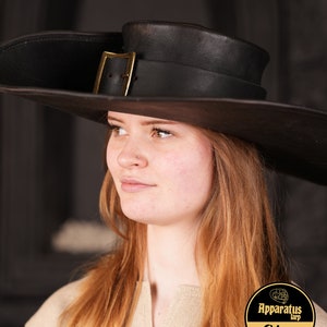 May include: A wide-brimmed black leather hat with a gold buckle. The hat is worn by a person with long, light brown hair. The person is wearing a light brown shirt. The image includes the text "Apparatus Larp Shop".
