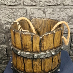 Könnte beinhalten: Ein rustikaler Holzeimer mit Metallbändern und Seilgriffen. Der Eimer hat ein verwittertes Aussehen mit vertikalen Holzlatten und ist so gestaltet, dass er einem alten Fass ähnelt. Der Eimer ist ein dekorativer Gegenstand.