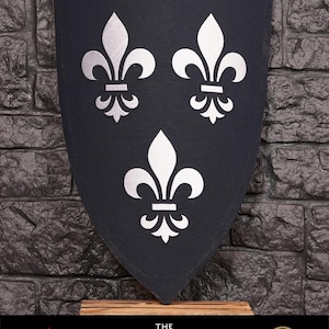 Op de afbeelding: Een zwart schild met drie zilveren lelies, een symbool van koninklijkheid. Het schild wordt getoond op een houten standaard. De tekst "The Witcher" is onder het schild gedrukt.