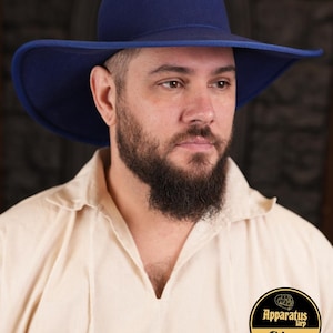 Puede incluir: Un hombre con un sombrero de fieltro azul cobalto de ala ancha. El sombrero tiene una corona ligeramente redondeada y un ala ancha y plana. El hombre lleva una camisa blanca con cuello. El texto "Apparatus Larp Shop" es visible en la esquina inferior derecha de la imagen.