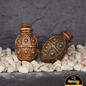 Könnte beinhalten: Zwei goldfarbene, runde Dekorationsflaschen mit Korkstopfen. Die Flaschen haben eine strukturierte Oberfläche mit einem geometrischen Muster und stehen auf einem Bett aus weißen Kieselsteinen.