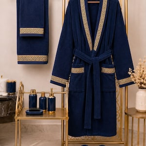 Puede incluir: Una bata de baño azul marino con un patrón griego dorado en el cuello, los puños y los bolsillos, acompañada de una toalla a juego, un par de pantuflas y un juego de accesorios de baño azul marino con detalles dorados sobre un soporte.