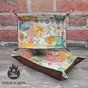 May include: Two rectangular valet trays with brown exterior sides and snap-button corners. The interior features a colourful patchwork pattern with floral and geometric motifs in shades of yellow, pink, teal, and red. The text GOBLIN AND WYRM is visible.
