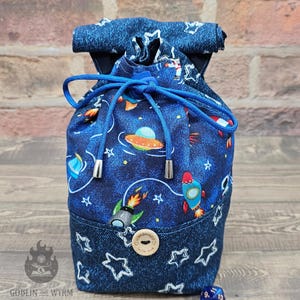 May include: A blue drawstring bag with a space-themed design of rockets, planets, and stars. The bag has a denim-style trim and a wooden button. The blue drawstring secures the bag, which is designed to hold small items.
