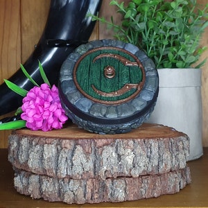 May include: A round, dark green and brown decorative box with a stone-like texture. The box has a circular opening with a green door and a metal handle. The box is sitting on a wooden surface.