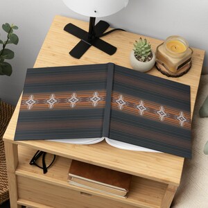May include: Open notebook with a dark gray cover featuring horizontal brown stripes and diamond-shaped patterns. The notebook is open on a light wood nightstand, next to a small succulent, a lit candle, and a lamp.