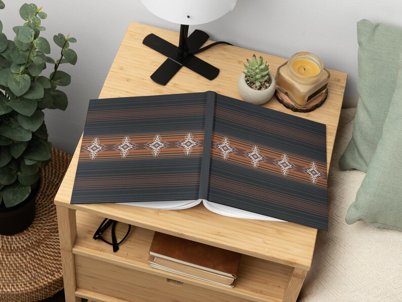 May include: Open notebook with a dark gray cover featuring horizontal brown stripes and diamond-shaped patterns. The notebook is open on a light wood nightstand, next to a small succulent, a lit candle, and a lamp.