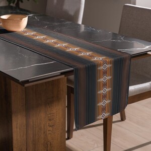 May include: A rectangular table runner with a striped pattern in black, brown, and orange. The runner features a repeating geometric design in the center. The runner is placed on a dark marble table, with chairs and a wooden table leg visible.