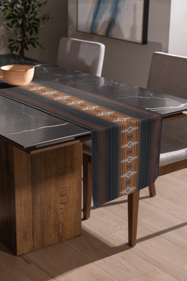 May include: A rectangular table runner with a striped pattern in black, brown, and orange. The runner features a repeating geometric design in the center. The runner is placed on a dark marble table, with chairs and a wooden table leg visible.