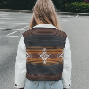May include: A white denim jacket with a patterned back panel. The back panel features a geometric design in shades of brown, black, and orange. The jacket has a classic collar and buttoned cuffs. The person is wearing blue jeans.