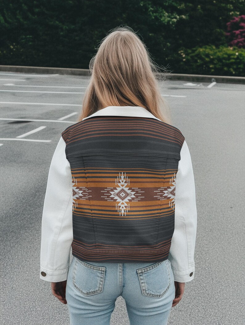 May include: A white denim jacket with a patterned back panel. The back panel features a geometric design in shades of brown, black, and orange. The jacket has a classic collar and buttoned cuffs. The person is wearing blue jeans.