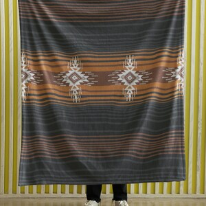 May include: A large, rectangular blanket with a geometric pattern. The blanket features horizontal stripes in shades of gray, brown, and orange. The design includes repeating diamond-shaped motifs in white and brown. The blanket is held up by a person.