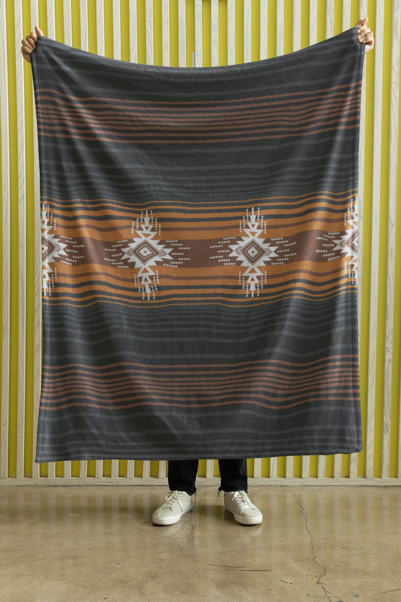 May include: A large, rectangular blanket with a geometric pattern. The blanket features horizontal stripes in shades of gray, brown, and orange. The design includes repeating diamond-shaped motifs in white and brown. The blanket is held up by a person.