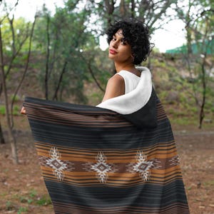 May include: A hooded blanket with a black, brown, and tan striped pattern and geometric designs. The blanket has a white, fluffy interior and is being held up in an outdoor setting.