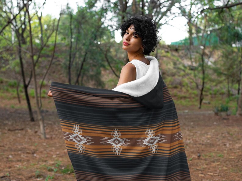 May include: A hooded blanket with a black, brown, and tan striped pattern and geometric designs. The blanket has a white, fluffy interior and is being held up in an outdoor setting.