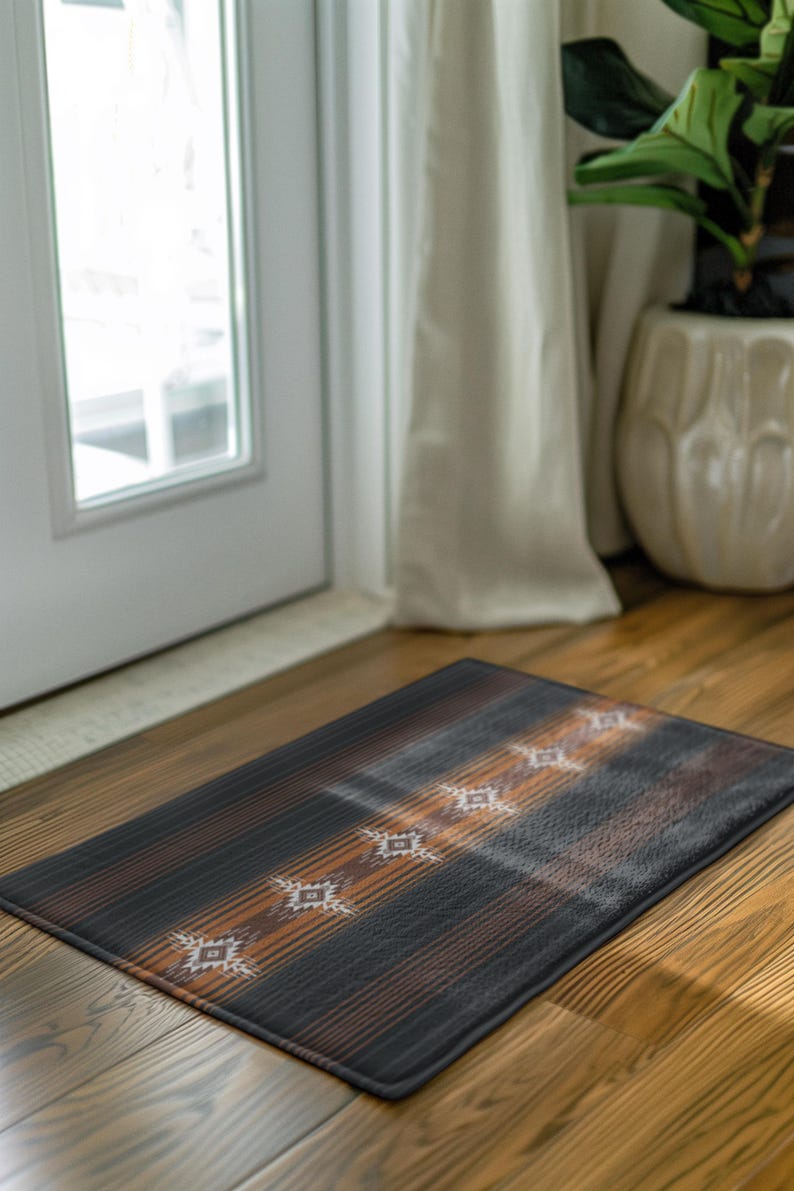 May include: Rectangular doormat with a dark brown and black striped pattern. The mat features a series of white geometric designs. The mat is placed on a wooden floor near a white door and a potted plant.