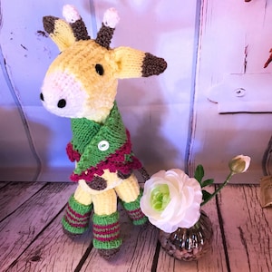 May include: A knitted giraffe toy wearing a green and pink scarf with a white button. The giraffe is standing on a wooden surface with a white flower in a glass vase.