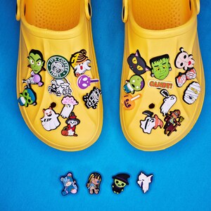 Könnte beinhalten: Zwei Paar gelbe Clogs, die mit Halloween-Themen-Anhängern verziert sind. Die Anhänger zeigen Geister, Frankensteins Monster, Fledermäuse und das Wort "CANDY!". Zusätzliche Anhänger sind darunter auf einer blauen Oberfläche verstreut.