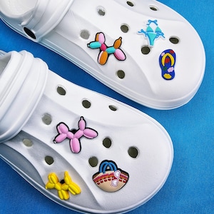 Könnte beinhalten: Zwei weiße Clogs, verziert mit bunten Charms. Ein Clog zeigt einen Ballonhund-Charm in Orange, Rosa und Blau, einen blauen Bikini-Charm und einen Flip-Flop-Charm. Der andere Clog hat einen rosa Ballonhund-Charm, einen gelben Ballonhund-Charm und einen Strandtaschen-Charm.