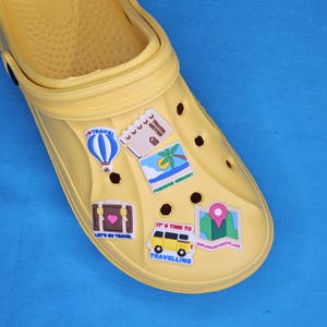 May include: Yellow clog shoe adorned with travel-themed charms. Charms include a hot air balloon, a suitcase with a heart, a notebook, a palm tree, a map, and a yellow van with the text "IT'S TIME TO TRAVELLING".