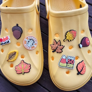 May include: Two pairs of yellow clogs adorned with fall-themed charms. Charms include an acorn, maple leaves, a slice of ham, and a Starbucks Pumpkin Spice logo. One charm reads "Me So Corny".