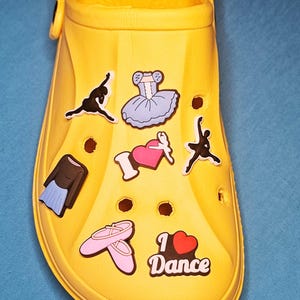Puede incluir: Zueco amarillo decorado con abalorios temáticos de ballet. Los abalorios incluyen bailarines de ballet, un tutú, zapatillas de ballet y la frase "I ❤️ Dance". El zapato está sobre una superficie azul.