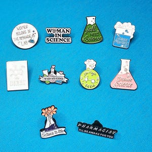 May include: A selection of enamel pins with science-related designs. The pins display phrases such as "Woman in Science," "I'm Very Into Science," and "Because Science." Other pins show lab equipment and the word "Chocolate."