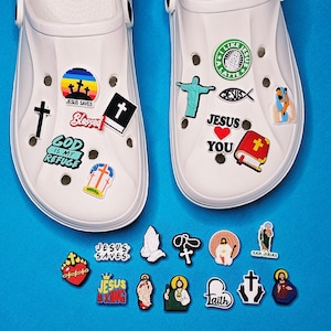 May include: White clogs adorned with religious charms. The charms feature crosses, the text "Jesus Saves," and other Christian symbols. The clogs are displayed on a blue surface, highlighting the colourful charms.