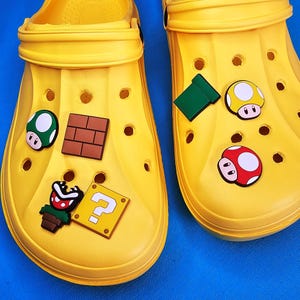 Puede incluir: Un par de zuecos amarillos brillantes decorados con coloridos dijes. Los dijes incluyen un hongo verde y blanco, un patrón de ladrillos marrones, un signo de interrogación, una tubería verde y hongos rojos y amarillos. Los zuecos están sobre una superficie azul.