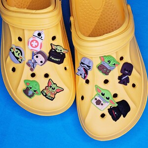 May include: Two pairs of yellow clogs decorated with Star Wars character charms. The charms feature Baby Yoda, BB-8, and Stormtroopers. The clogs are displayed on a bright blue surface.