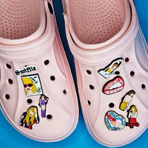 Puede incluir: Un par de Crocs de color rosa claro adornados con varios dijes decorativos. Los dijes incluyen imágenes de una mujer rubia, el texto "Swiftie" y "1989", y otros gráficos. Los Crocs están sobre una superficie azul.