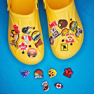 May include: Yellow clogs decorated with a variety of charms. The charms display images such as a kiwi, a maple leaf, and the text "eh?". Additional charms are placed below the clogs on a blue surface. These accessories are ideal for customising footwear.