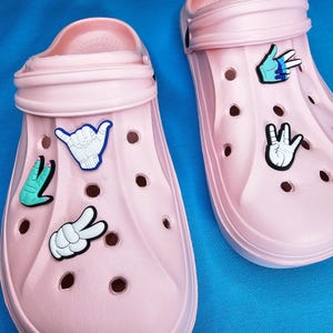 May include: Two pairs of pale pink clogs adorned with hand gesture charms. The charms depict a peace sign, a shaka, and an "okay" hand sign. The clogs are made of a rubbery material and have several ventilation holes.