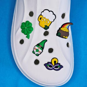 Op de afbeelding: Witte schoen versierd met kleurrijke bedels. Bedels zijn onder meer een groene klaver, een gele bierpul met wit schuim, een pot goud met een regenboog, een groene kabouter en een paars en geel masker. De schoen staat op een blauwe achtergrond.